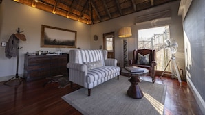 A room with a striped sofa, a wooden dresser, a standing lamp, a fan, and a large window with a view of a fence. - Evolve Back Kalahari (Hainaveld Game Farm)