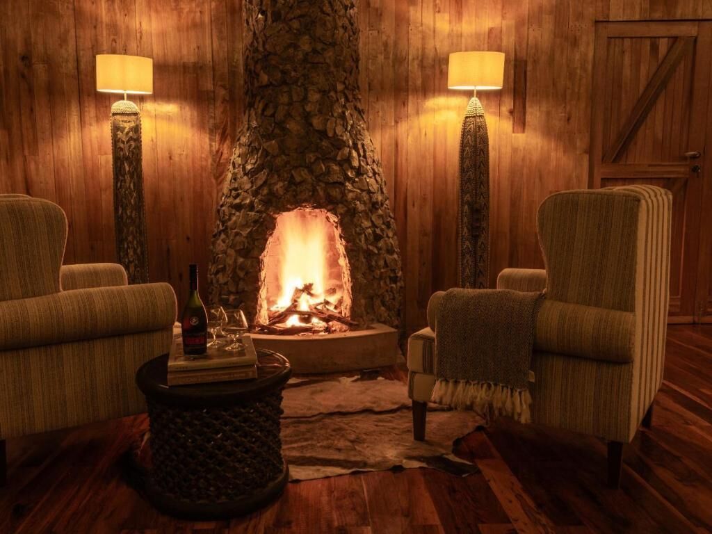 Una habitación acogedora con chimenea de piedra, dos sillones, una mesita con una botella y vasos, y una puerta de madera al fondo.