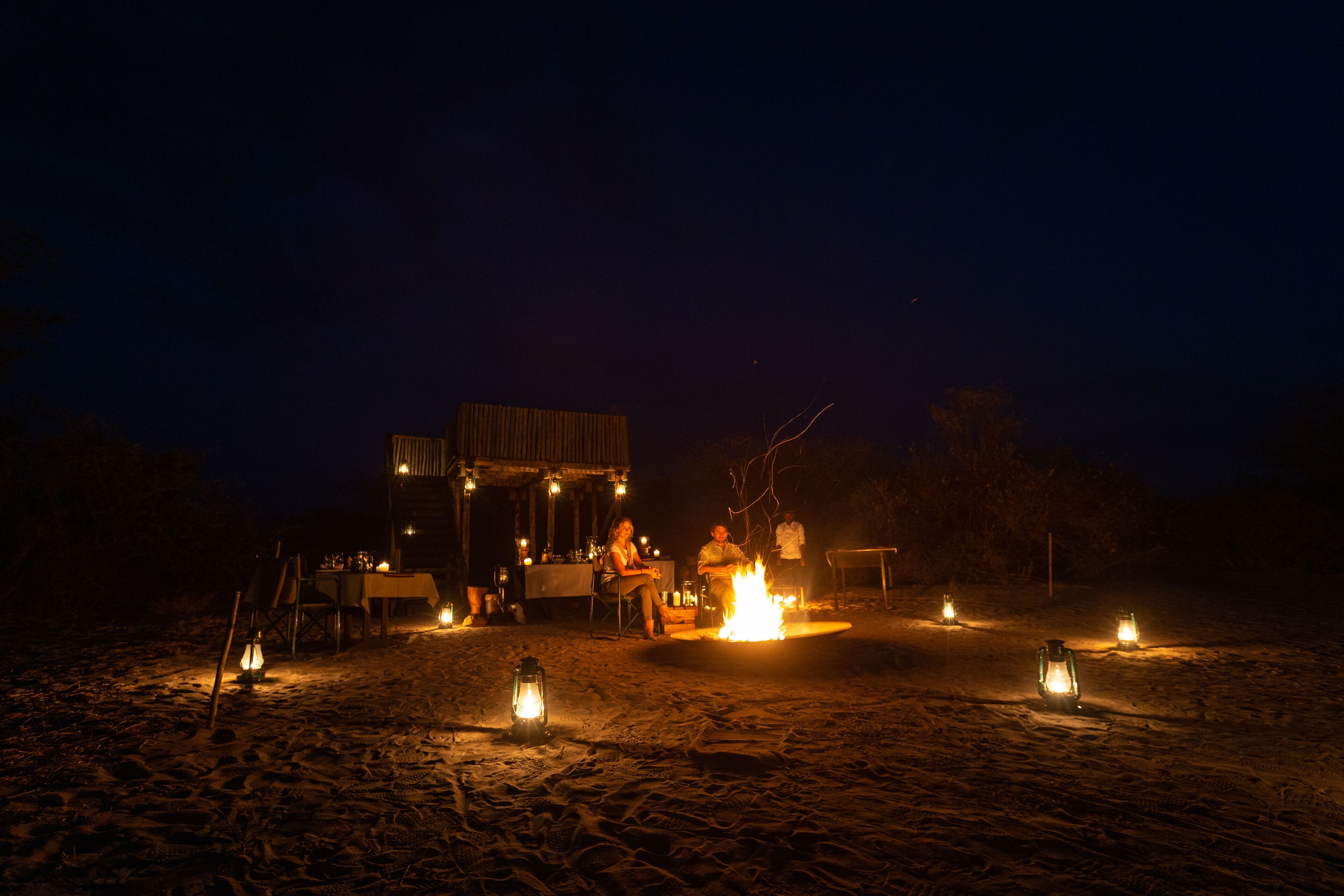 A beachside dining setup with a fire pit, tables, and chairs, surrounded by lanterns.
