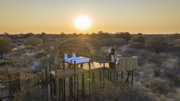 A wooden deck with a bed and a dining area, surrounded by a wooden fence and bushy vegetation. - Evolve Back Kalahari (Hainaveld Game Farm)