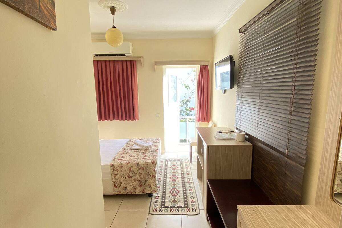 A compact hotel room with a bed, a small desk, a TV, and a window with blinds.