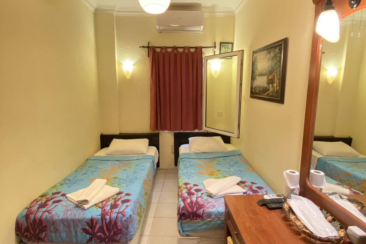 A hotel room with two beds, a wooden dresser, a mirror, a painting on the wall, and an air conditioning unit.