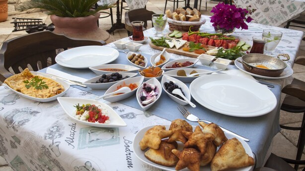 A table set with various dishes, including a plate of pastries, a bowl of olives, and a dish of tomatoes. - Alya Ruzgari Otel (Cesme)