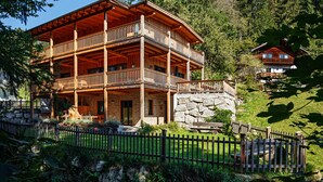 Una cabaña de madera de varios pisos con balcones, rodeada por un muro de piedra y una cerca de madera.