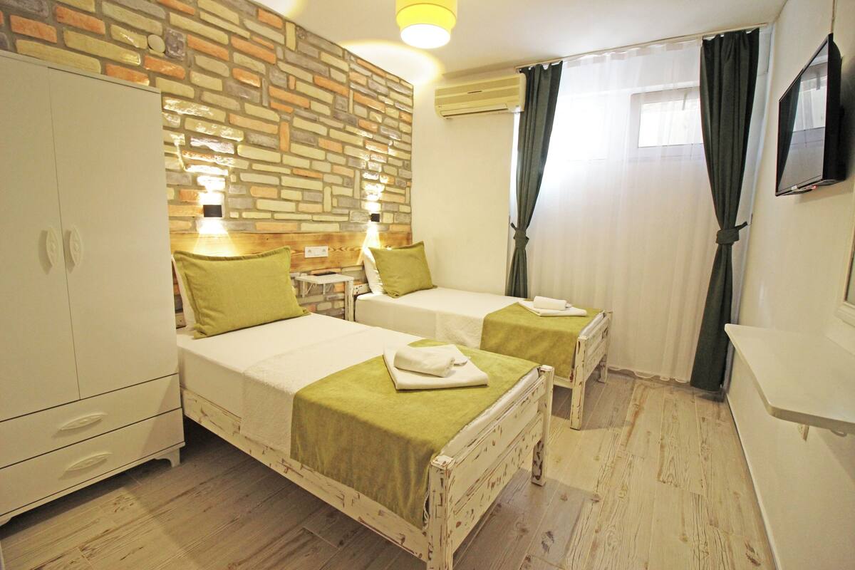 A hotel room with two beds, a brick-patterned wall, a wooden floor, a wardrobe, and a flat-screen TV.
