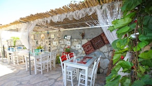 Un patio al aire libre con suelo de piedra, mesas y sillas blancas, y una pérgola con toldo blanco.