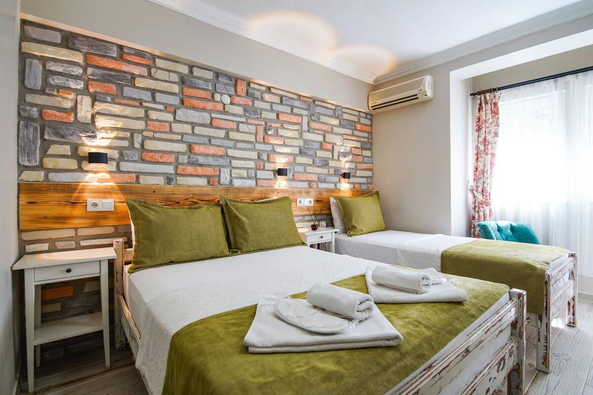 A hotel room with two beds, a stone wall, wooden headboard, and a window with curtains.