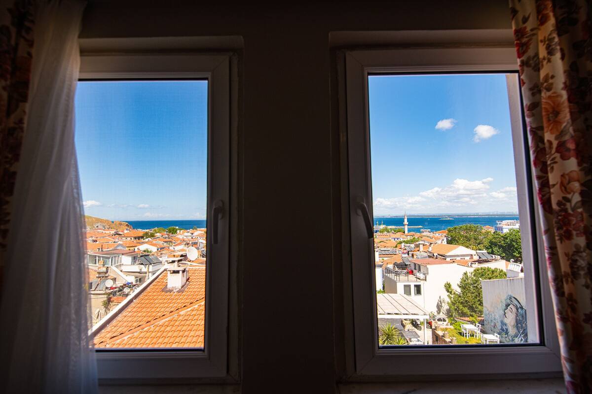 A view through two windows with curtains, overlooking a coastal town with buildings and a lighthouse.