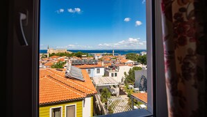 Vista desde una ventana que da a un pueblo con tejados rojos, una iglesia y un castillo lejano junto al mar.