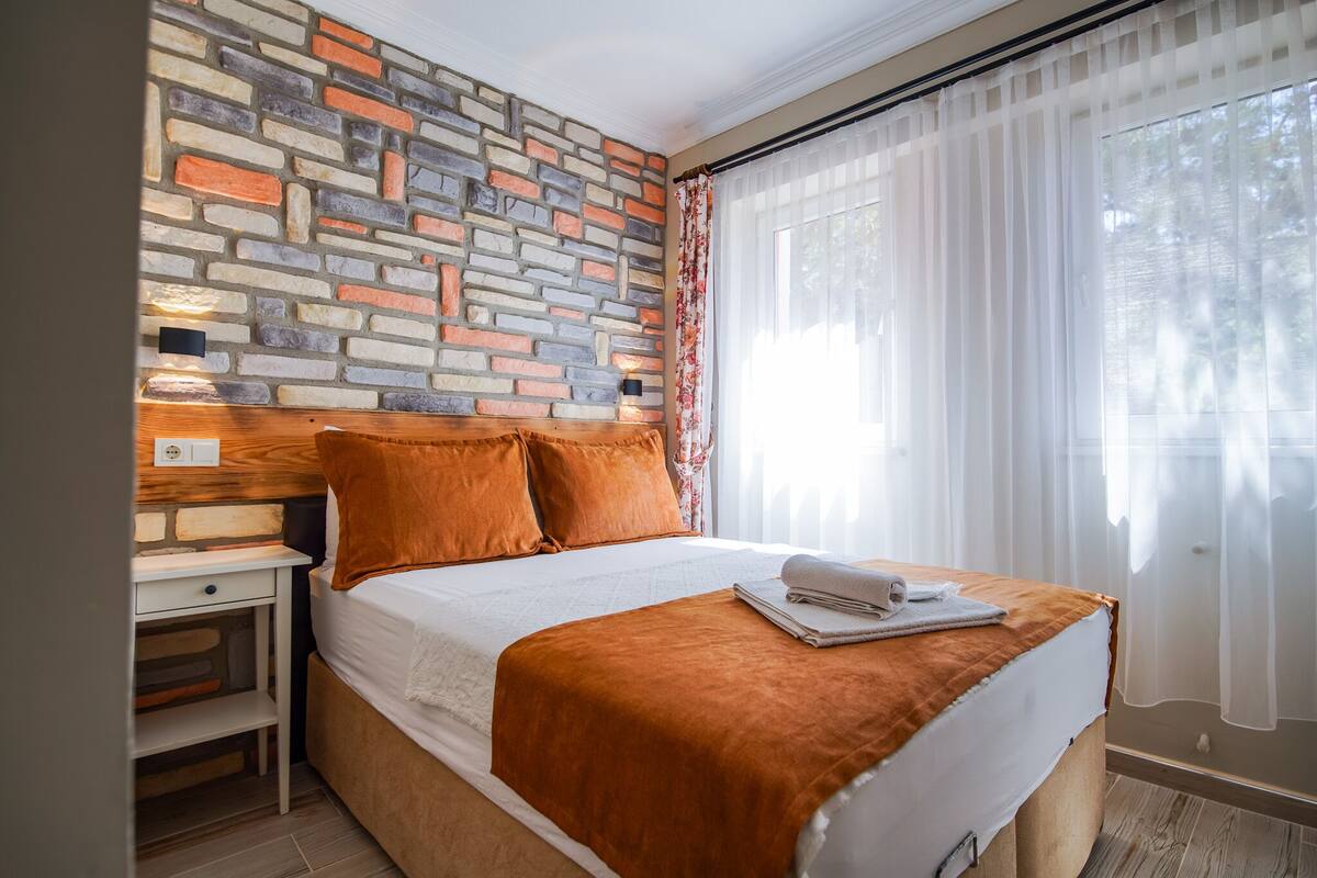 A neatly made bed with a brown blanket and pillows, a wooden headboard, a nightstand with a drawer, and a brick-patterned wall.