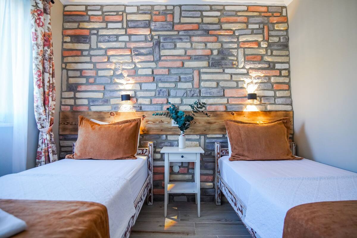 A room with two beds, a small table with a vase, and a stone wall.