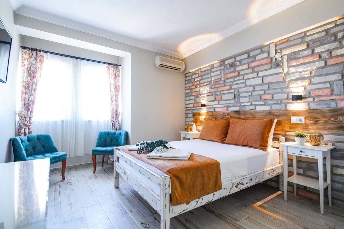 A bedroom with a white bed, two blue chairs, a wooden nightstand, and a brick-patterned wall.