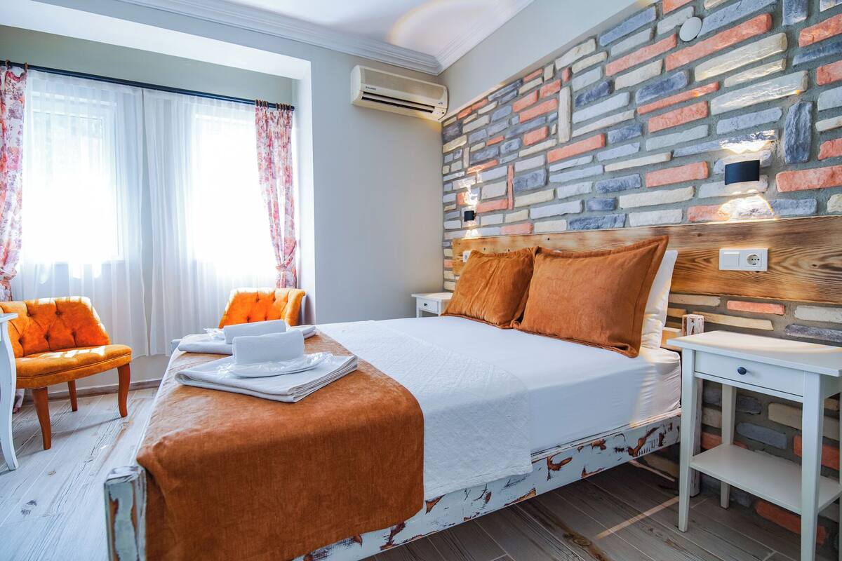 A modern hotel room with a brick-patterned headboard, a neatly made bed with orange bedding, a white bedside table, and a chair with orange cushions.