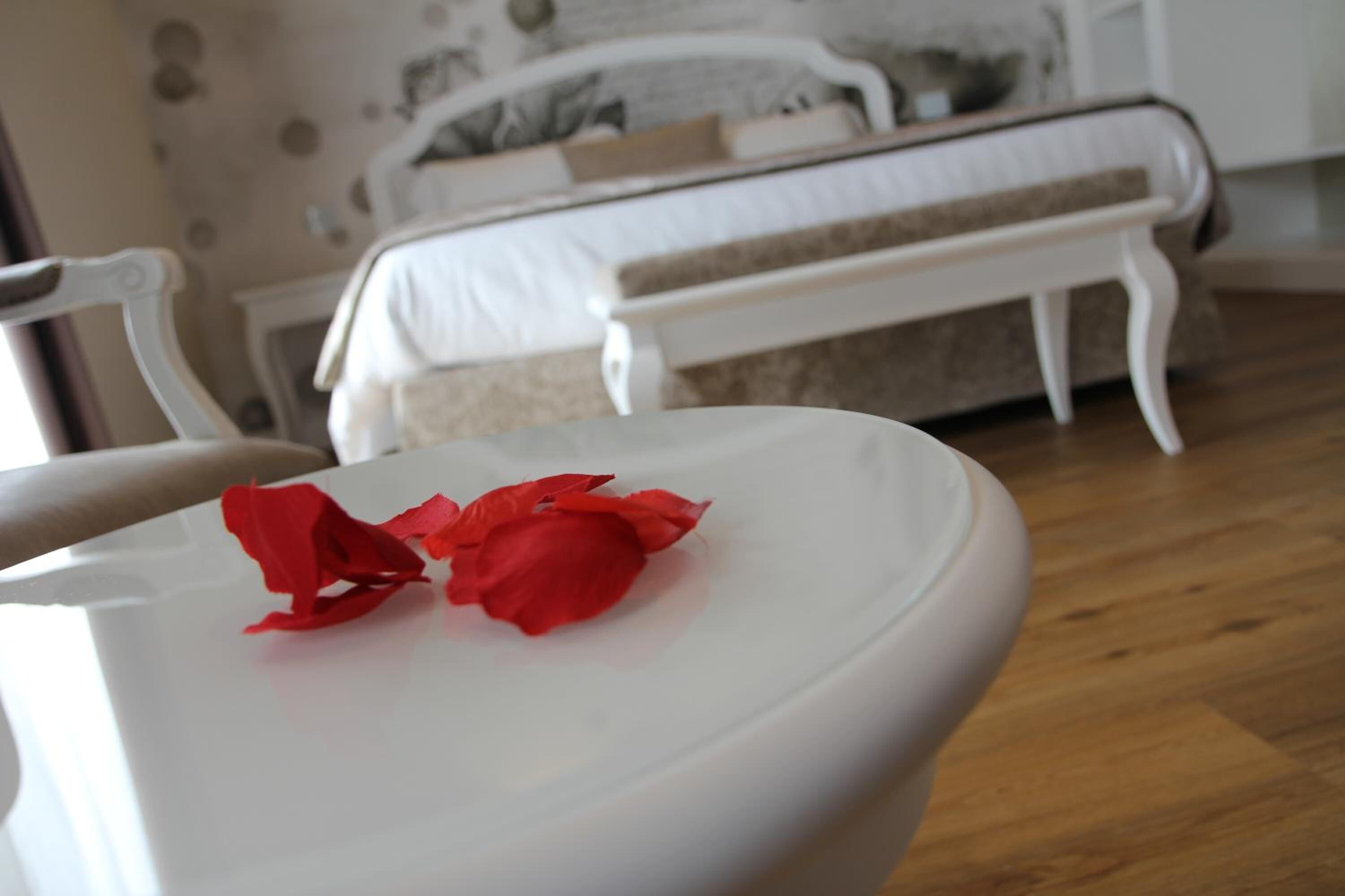 Una mesa blanca con pétalos de rosa roja en una habitación con cama y muebles blancos.