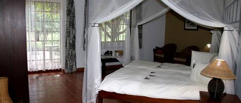 A bedroom with a four-poster bed, a bedside lamp, and a view of the outdoors through a window with sheer curtains.