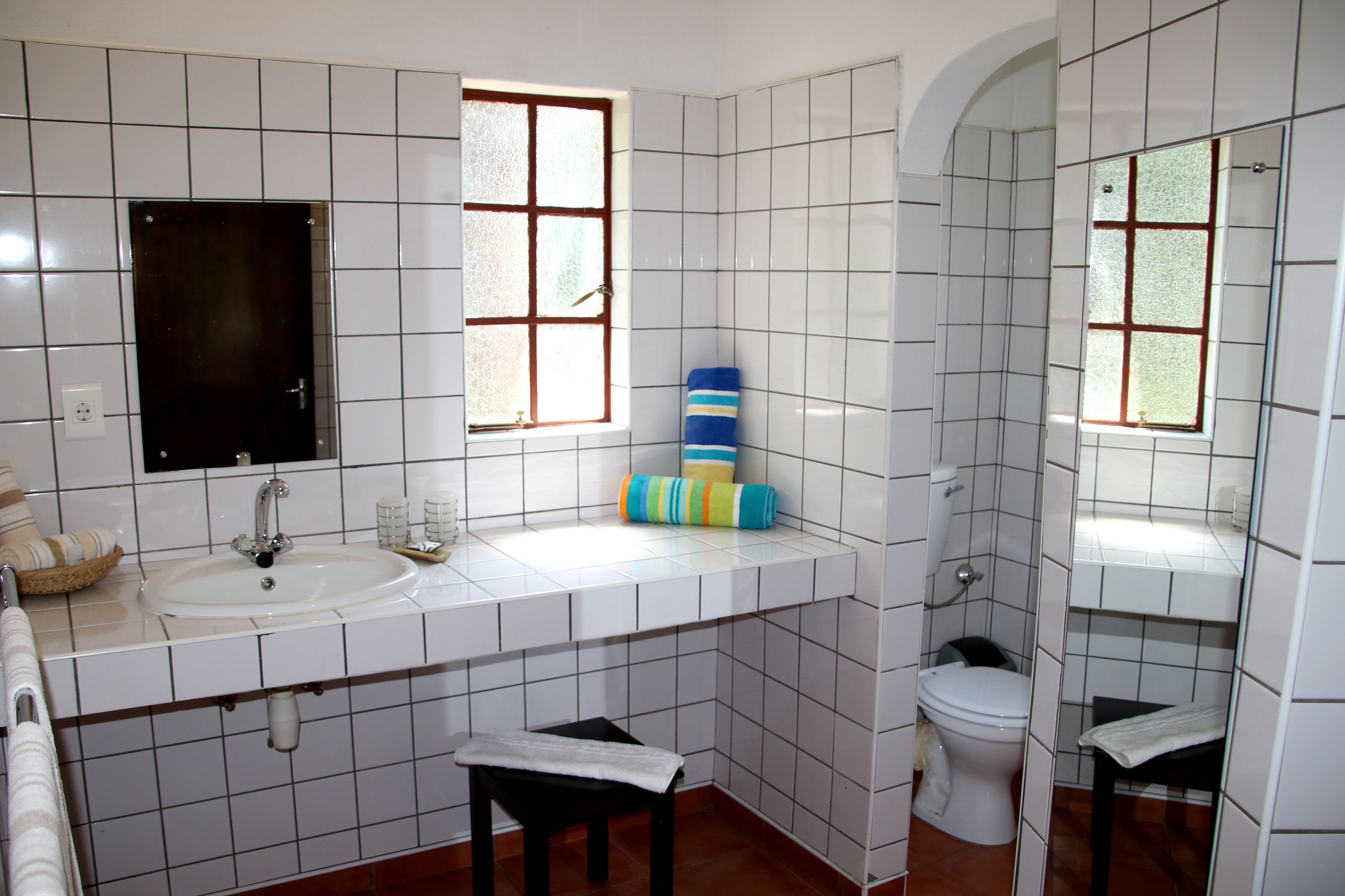 A bathroom with white tiled walls, a sink, a toilet, and two windows.