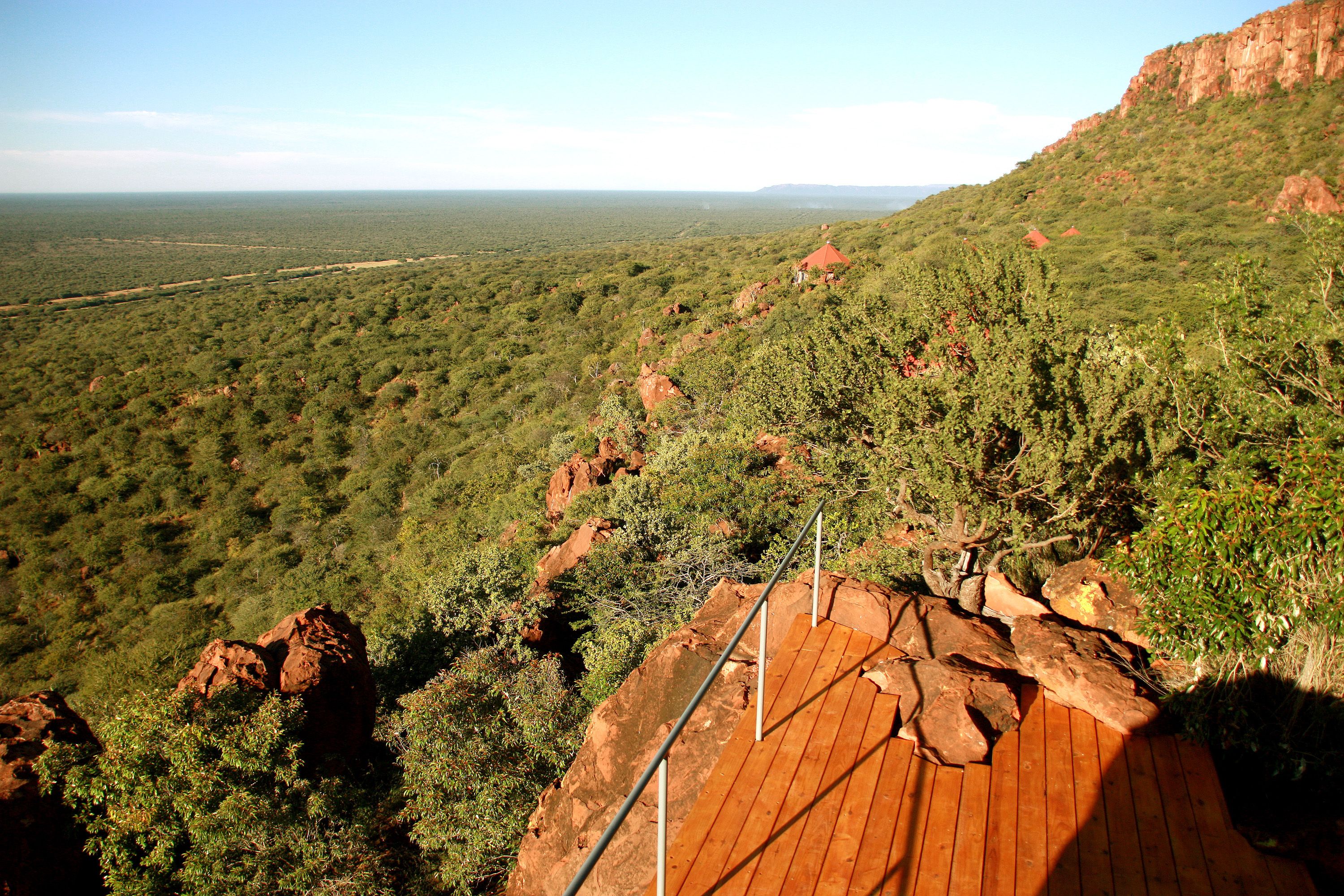 A wooden deck with metal railings overlooking a vast landscape with red rock formations and dense greenery.