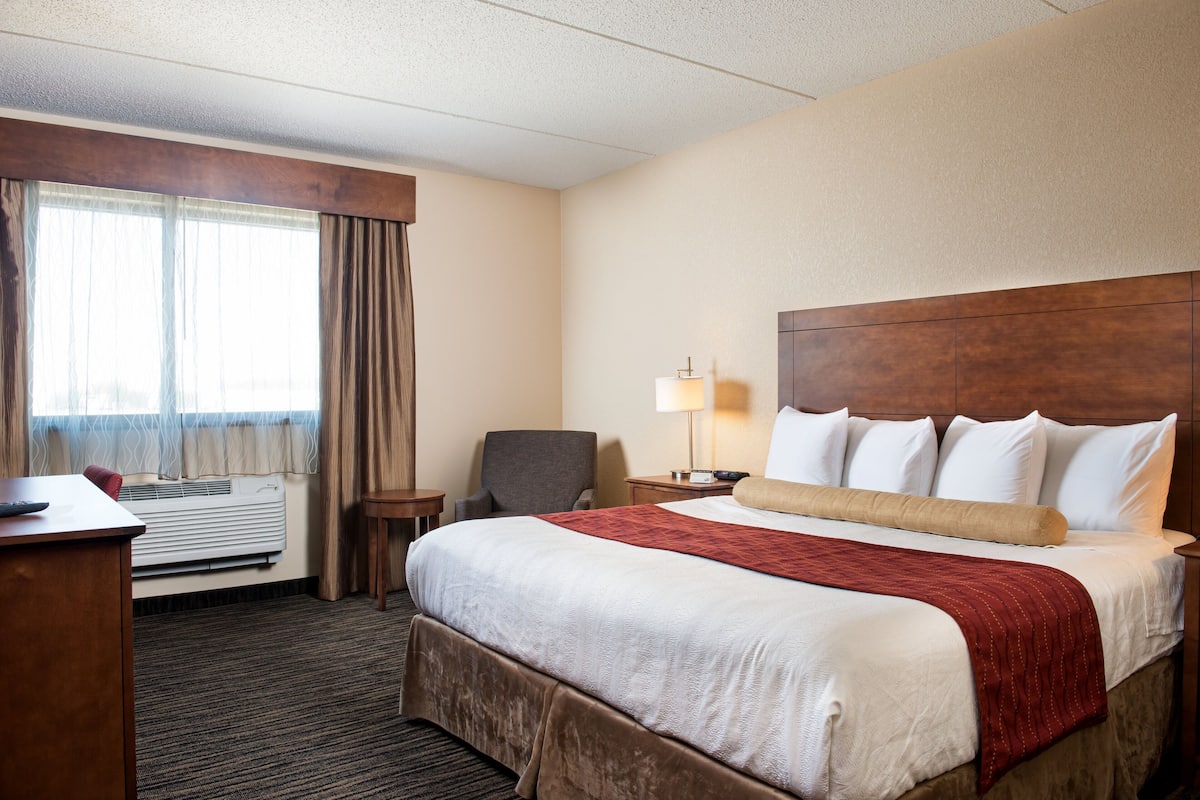 Ein Hotelzimmer mit einem großen Bett, einem Schreibtisch, einem Stuhl, einer Lampe und einem Vorhangfenster.