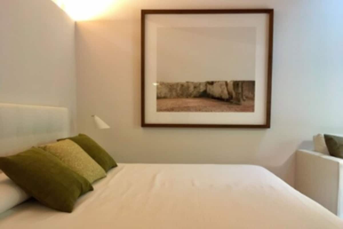 A neatly made bed with white bedding and green pillows, a framed picture on the wall, and a bedside table with a lamp.