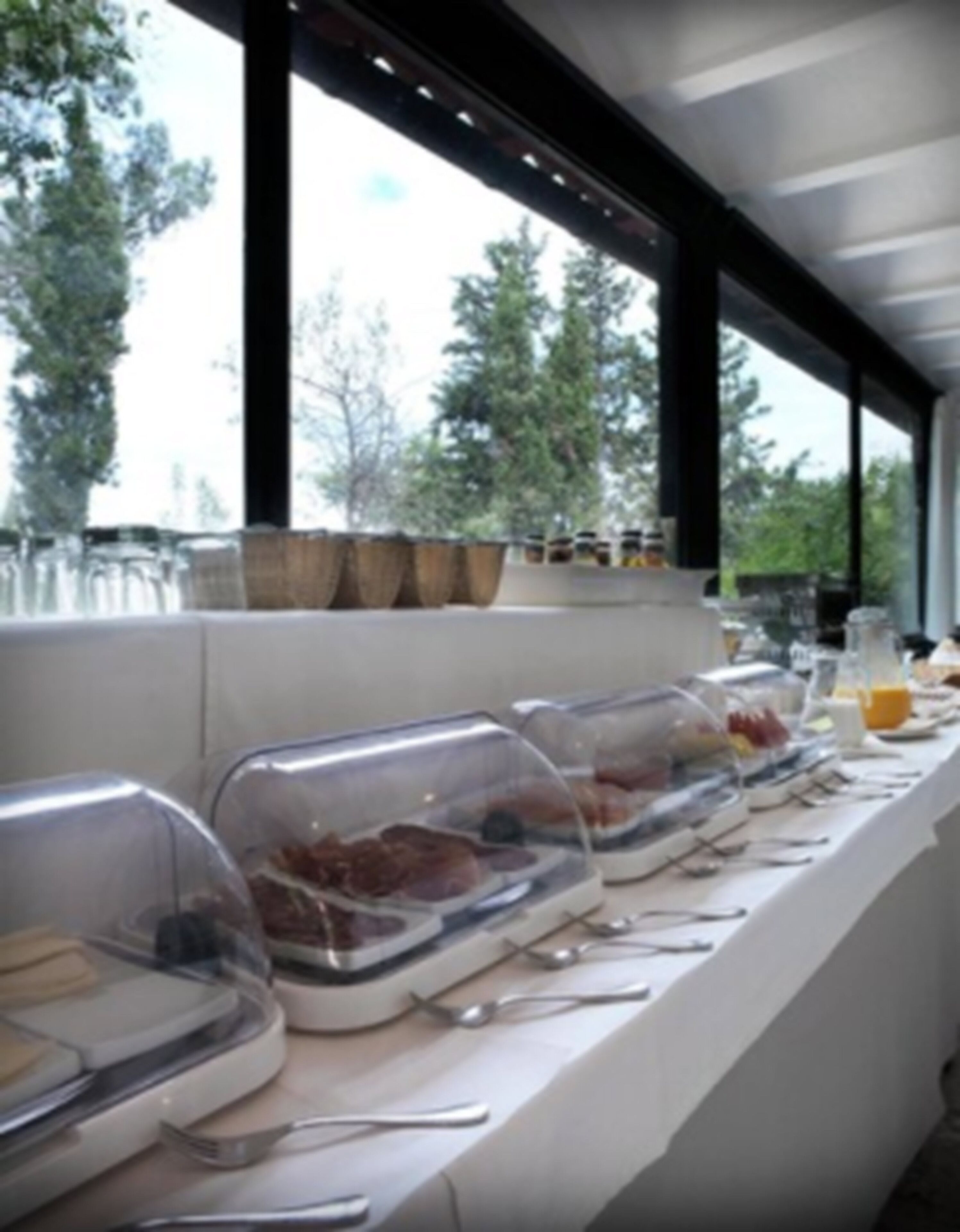 Montaje de bufé con recipientes cubiertos y utensilios sobre una mesa, con vista a árboles a través de amplias ventanas.