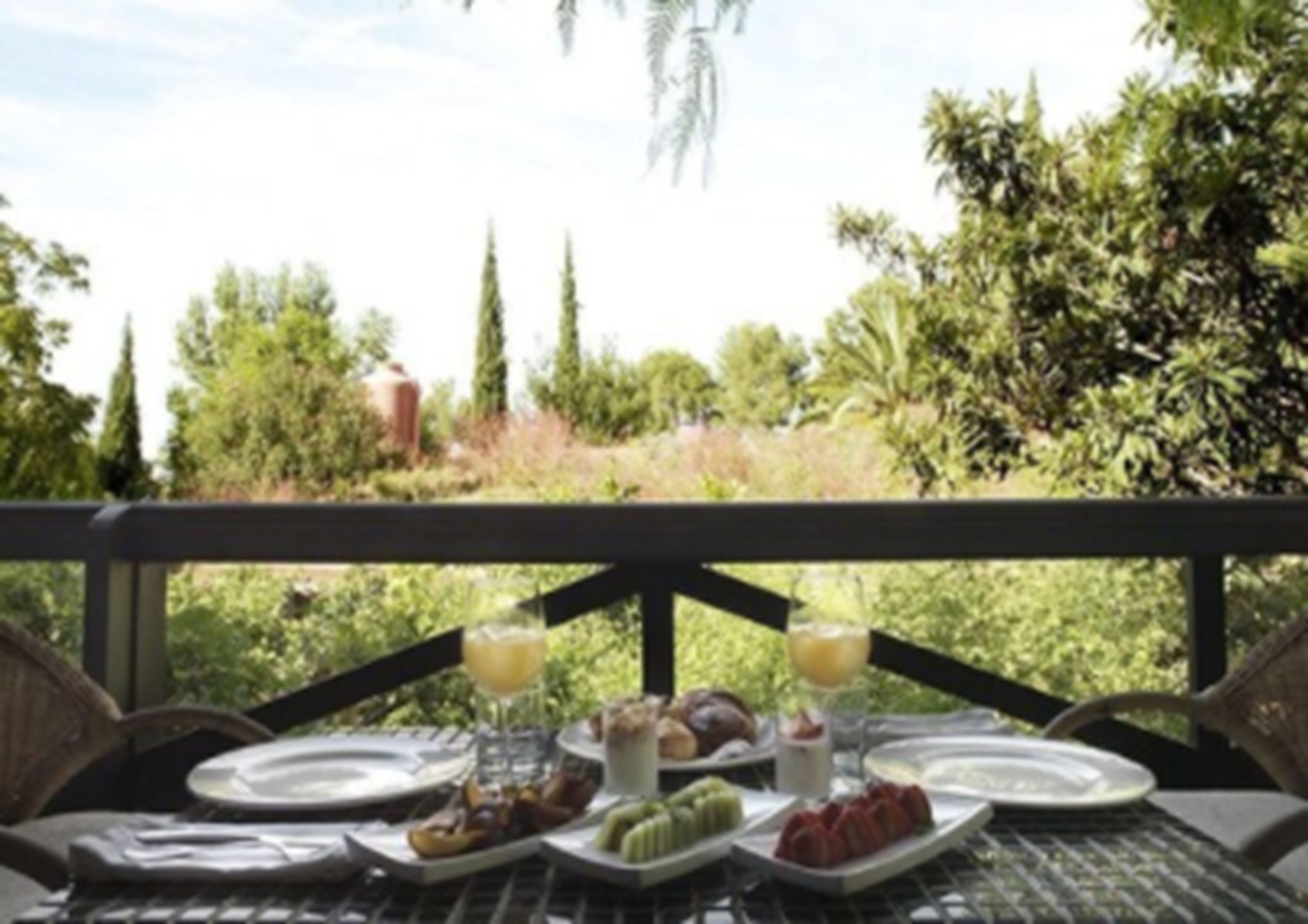 A table set outdoors with plates of food, glasses of juice, and a view of greenery and trees.