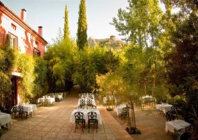 Un área de comedor al aire libre con mesas y sillas dispuestas sobre una superficie pavimentada, rodeada de árboles y un edificio en el fondo.