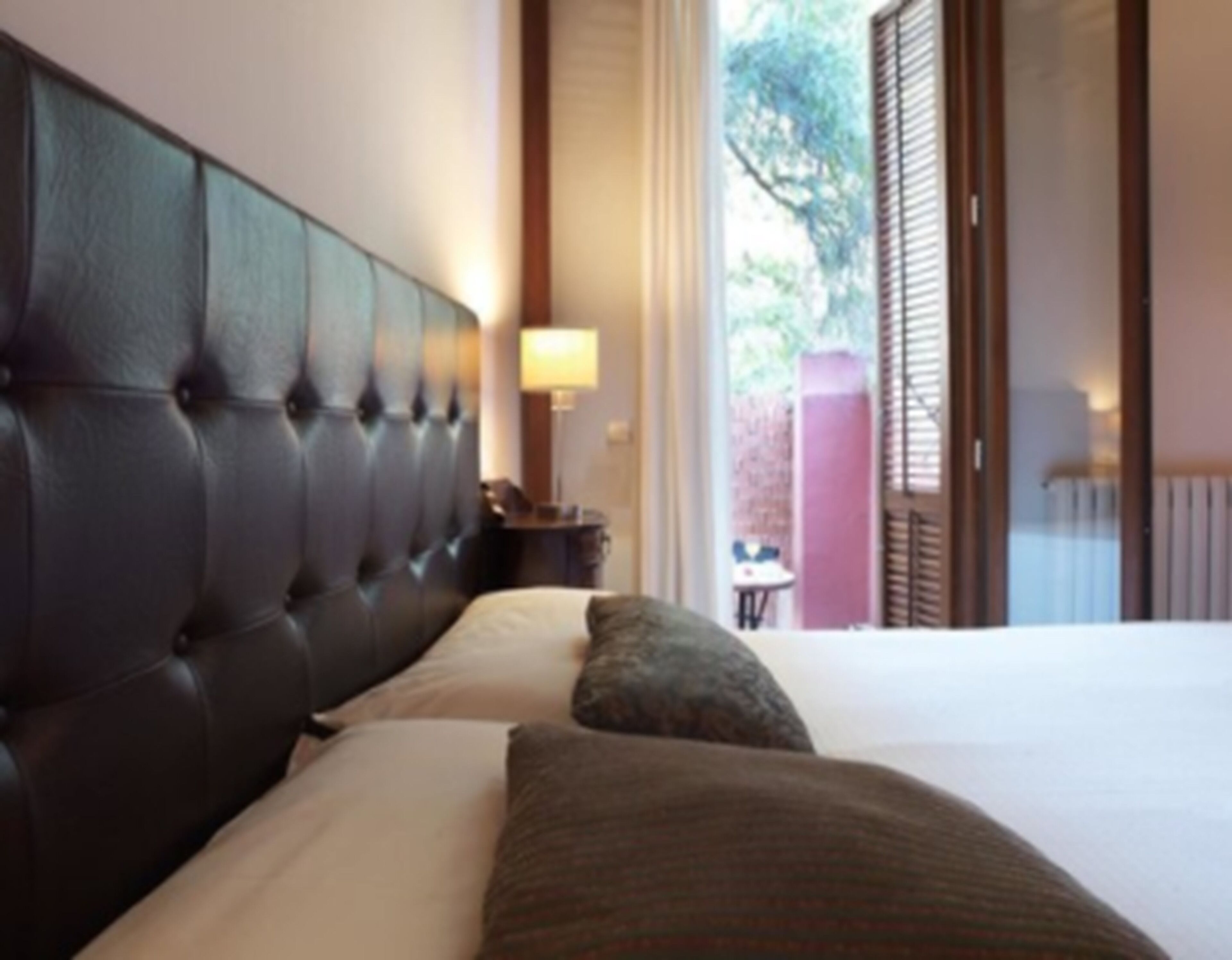 A hotel room with a tufted headboard, a bed with white linens, a nightstand with a lamp, and a view of the outdoors through a window with blinds.