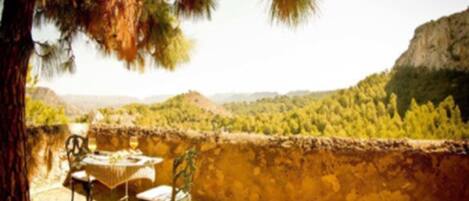 A patio with a table and chairs, overlooking a valley with greenery and a rocky cliff.