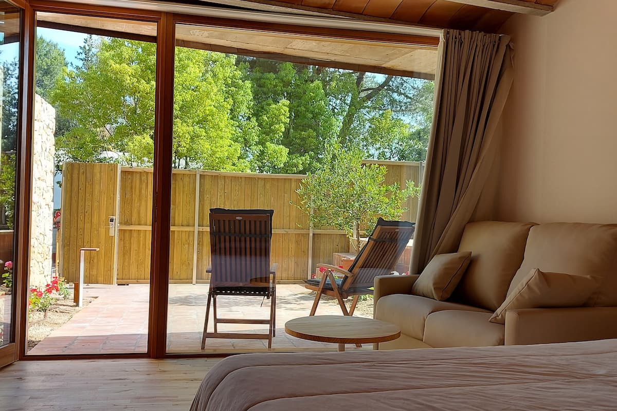A bedroom with a large bed, a sofa, and a chair. There is a sliding glass door leading to a patio with a wooden fence and a chair.
