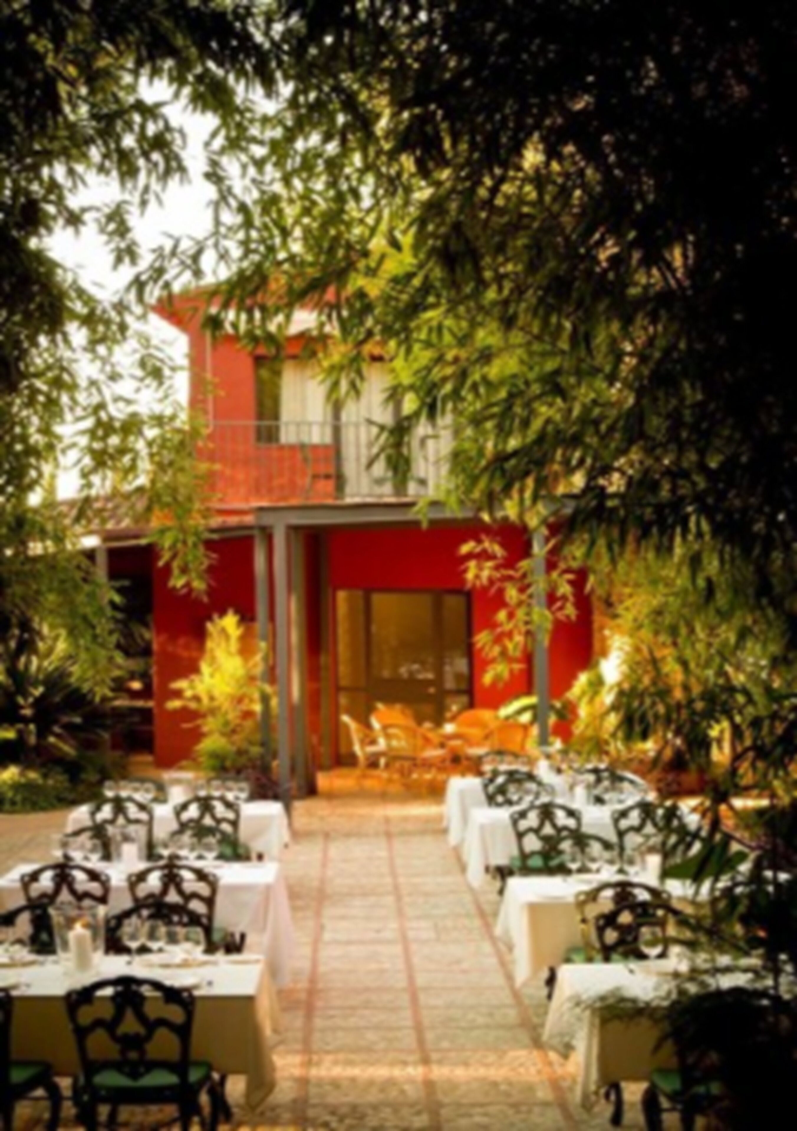 An outdoor dining area with tables and chairs, a red building in the background, and trees surrounding the space.