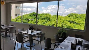 Un comedor moderno con mesas y sillas, ventanas amplias con vistas a zonas verdes y un edificio.