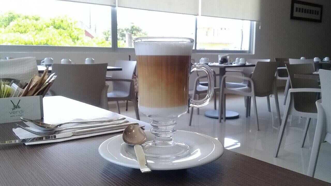 A clear glass mug with a layered beverage on a white plate, placed on a table in a bright dining area with white chairs and a view of greenery outside.