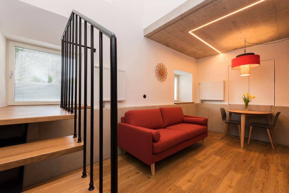 A modern living room with a red sofa, wooden flooring, a dining table with chairs, a staircase with black railings, and a pendant light.