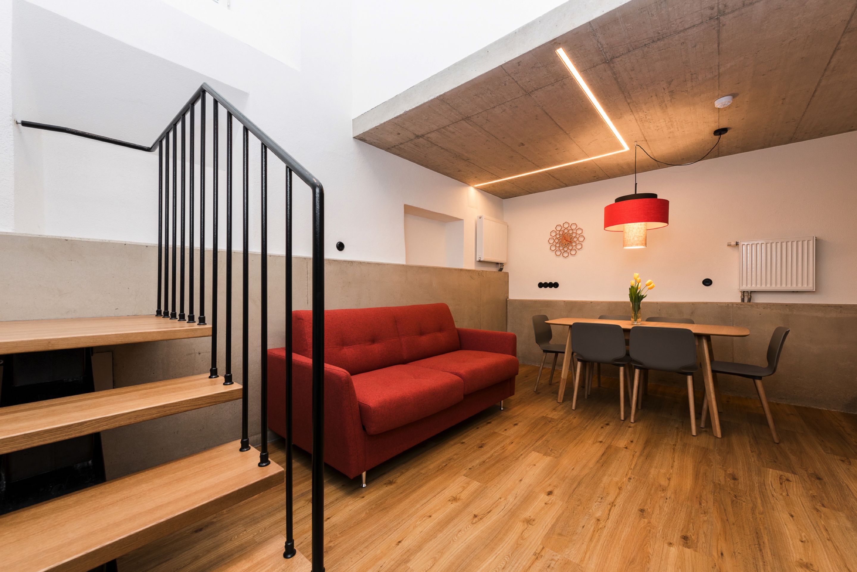 Un interior moderno con una escalera de madera, un sofá rojo, una mesa de comedor con sillas y una lámpara colgante.