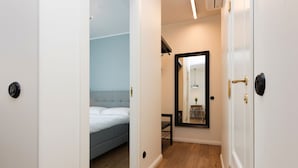 Un pasillo estrecho con piso de madera, una cama con cabecera gris, un espejo en la pared y una puerta con picaporte de latón.