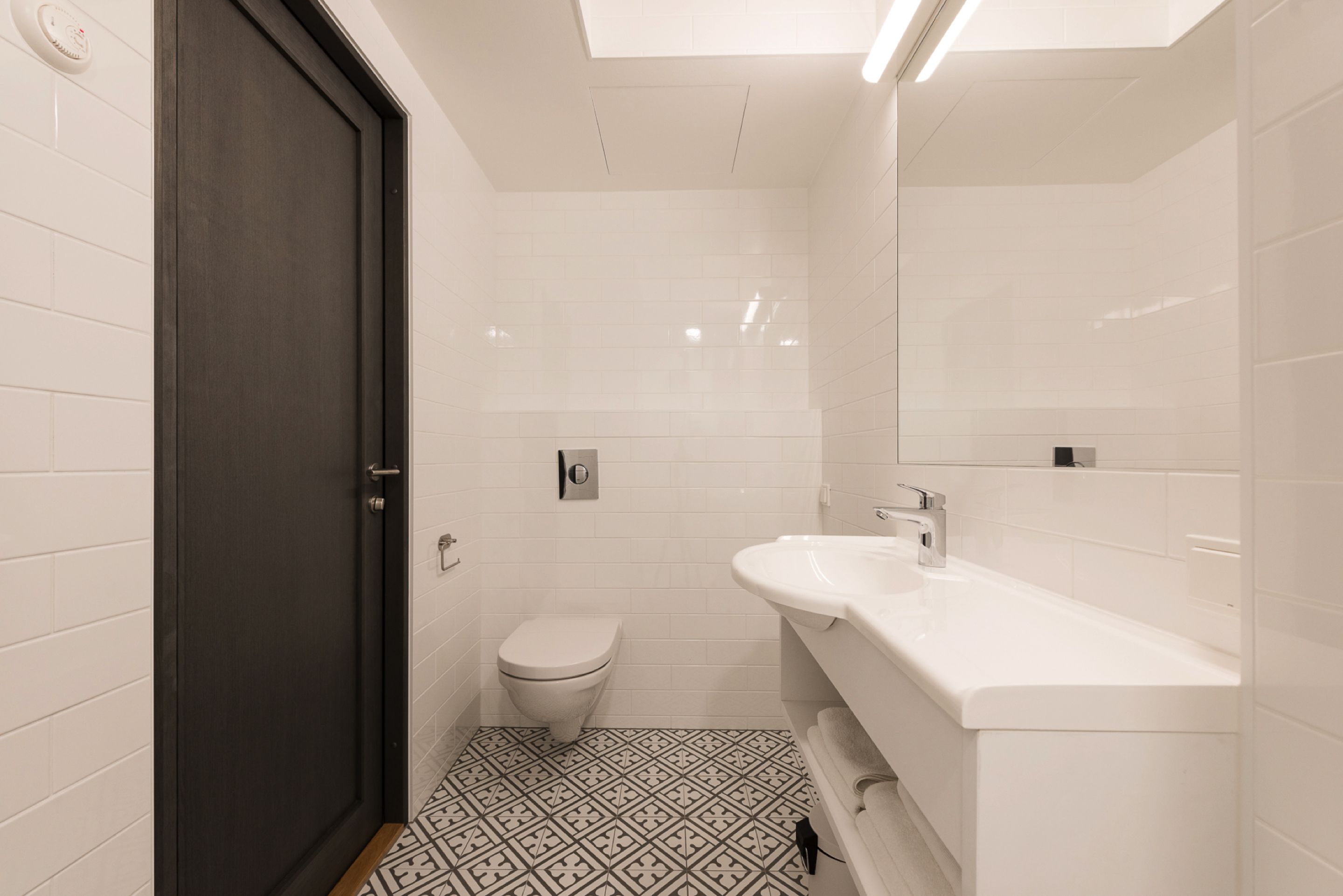 A modern bathroom with a dark door, white tiles, a toilet, and a white sink with a mirror above it.