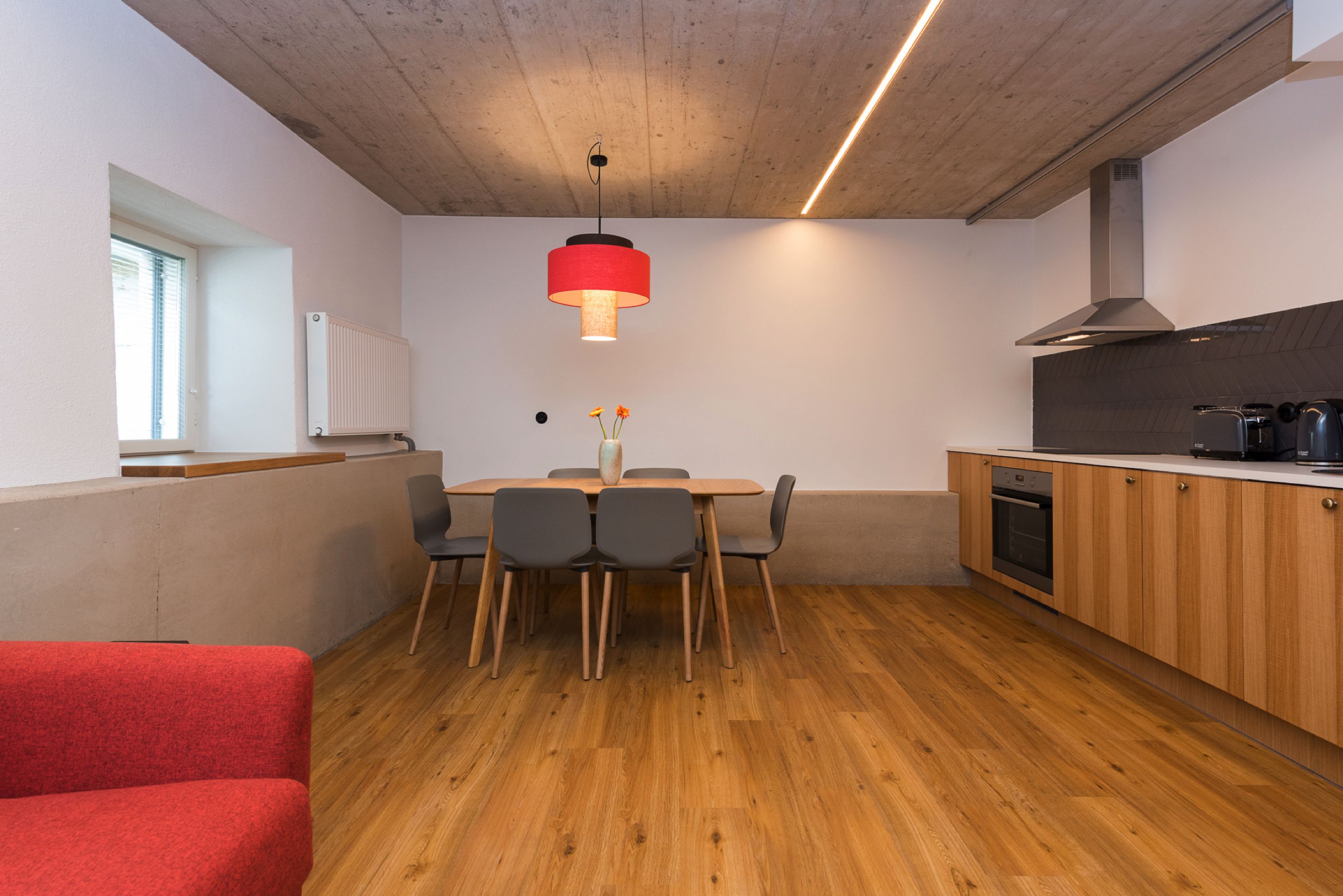 Une cuisine moderne avec des meubles en bois, un four encastré et une suspension rouge au-dessus de la table à manger.