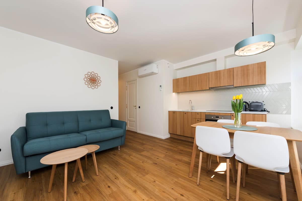 A modern apartment with a teal sofa, wooden dining table, and chairs. There is a kitchen area with wooden cabinets and a built-in oven.