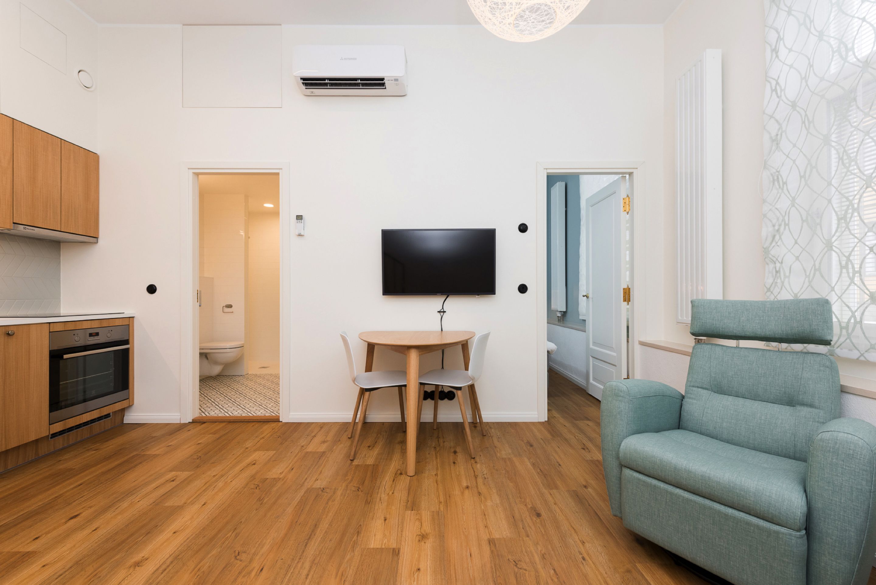 A modern apartment with a wooden floor, a dining table with chairs, a flat-screen TV mounted on the wall, a sofa, and a kitchen area with wooden cabinets and an oven.