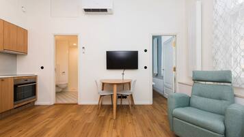 A modern apartment with a wooden floor, a dining table with chairs, a flat-screen TV mounted on the wall, a sofa, and a kitchen area with wooden cabinets and an oven.