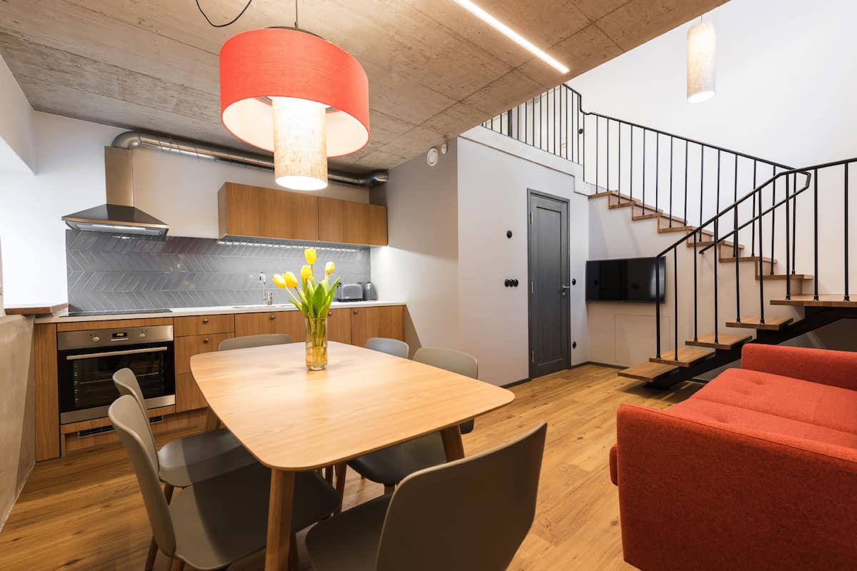 A modern kitchen with a wooden table and chairs, a red hanging light, and a staircase with a black railing.