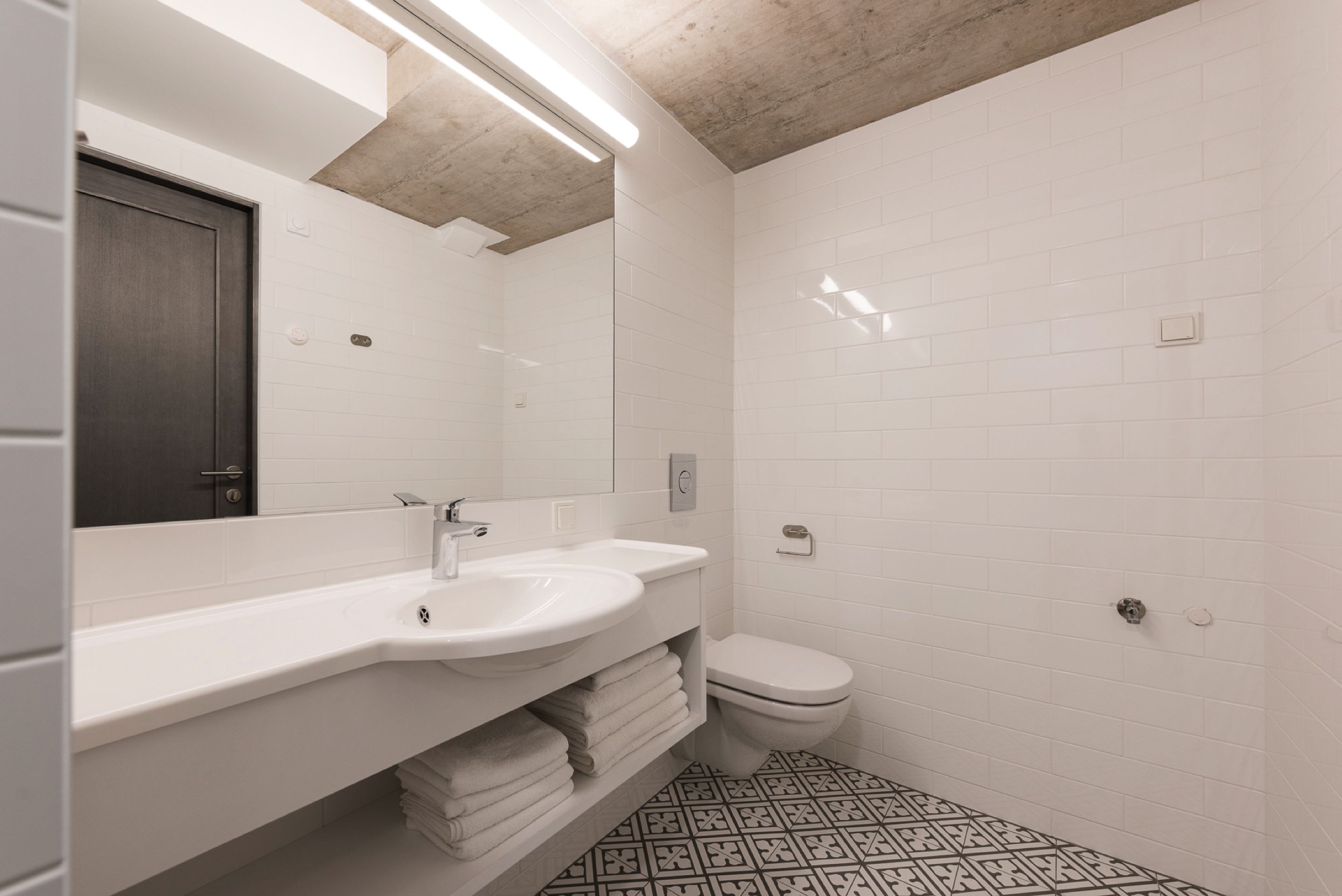 A modern bathroom with white tiles, a sink, a toilet, and a patterned floor.