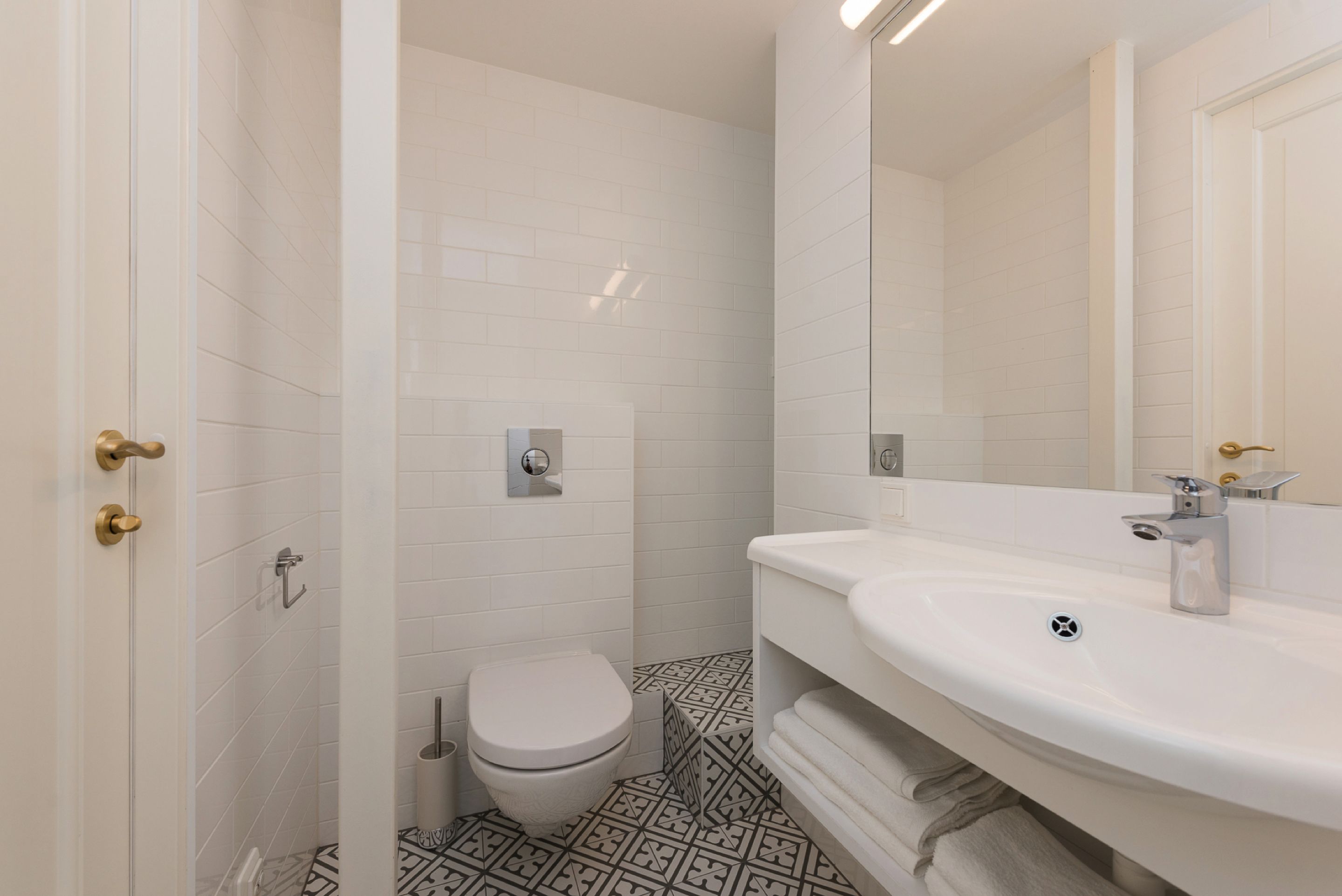 Un bagno moderno rivestito di piastrelle bianche, con un water, un lavandino con uno specchio e un pavimento decorato.
