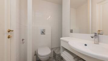 Un bagno moderno rivestito di piastrelle bianche, con un water, un lavandino con uno specchio e un pavimento decorato.