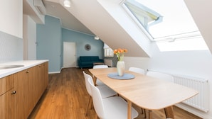 Una cocina moderna con armarios de madera, una encimera blanca y un comedor con mesa de madera y sillas blancas. Hay un tragaluz sobre el comedor y una pared azul al fondo.