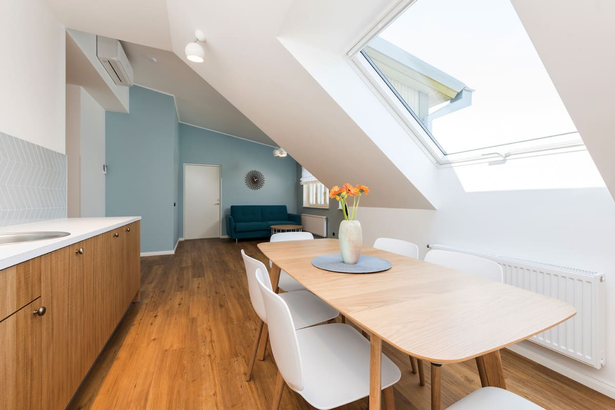 A modern kitchen with wooden cabinets, a white countertop, and a dining area with a wooden table and white chairs. There is a skylight above the dining area and a blue wall in the background.