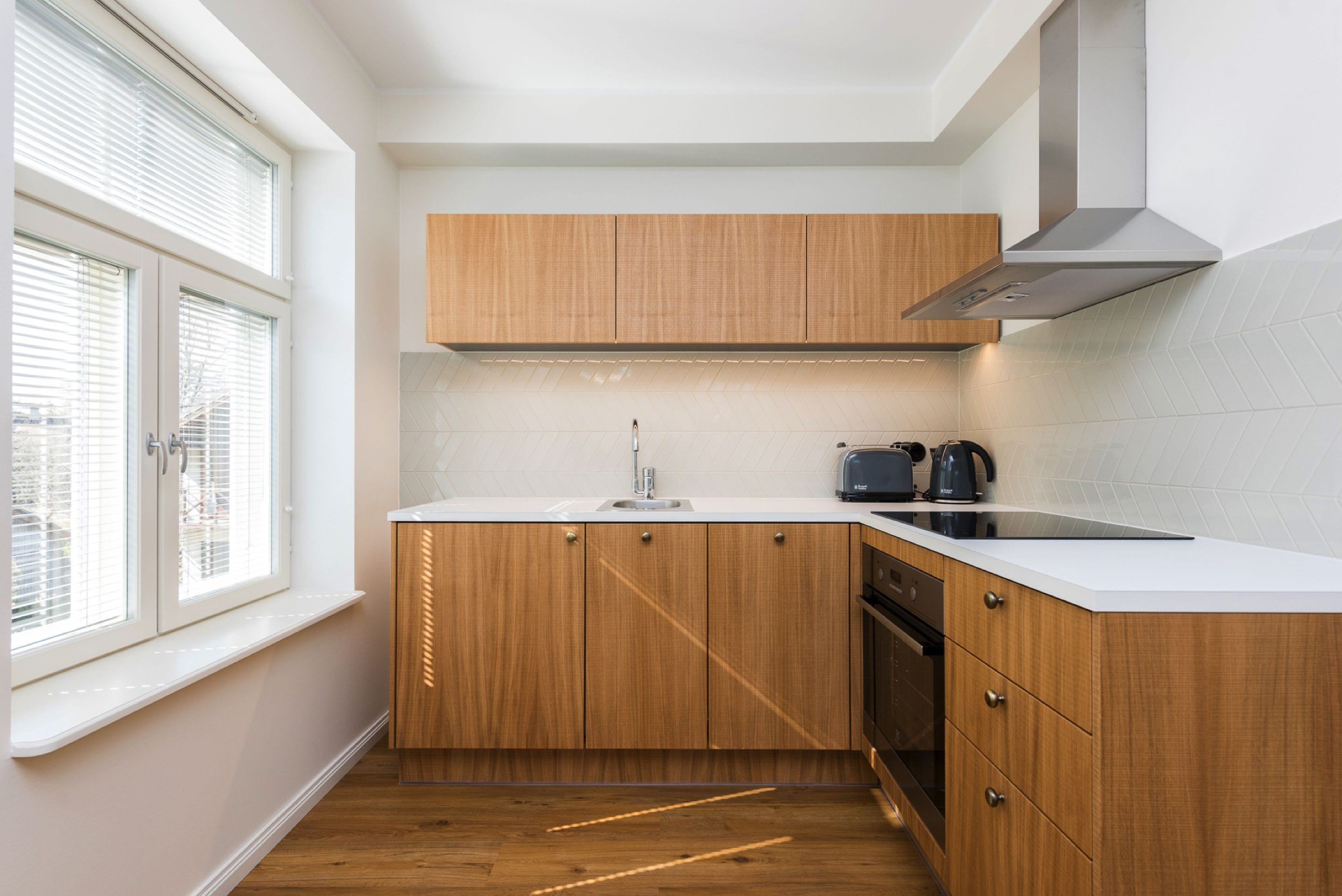 Una cocina compacta con armarios de madera, una encimera blanca, un extractor de acero inoxidable y una ventana con persianas.