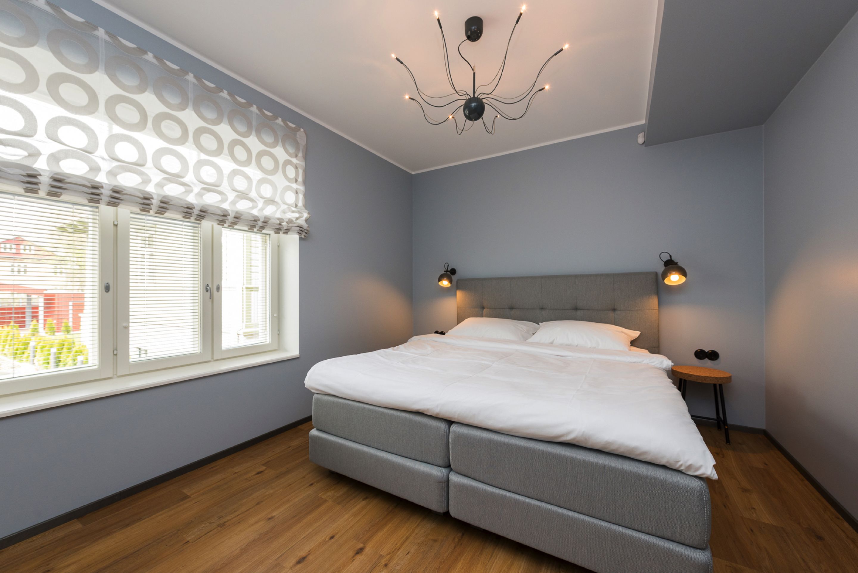 A bedroom with a bed, a window with blinds, a chandelier, and a nightstand.