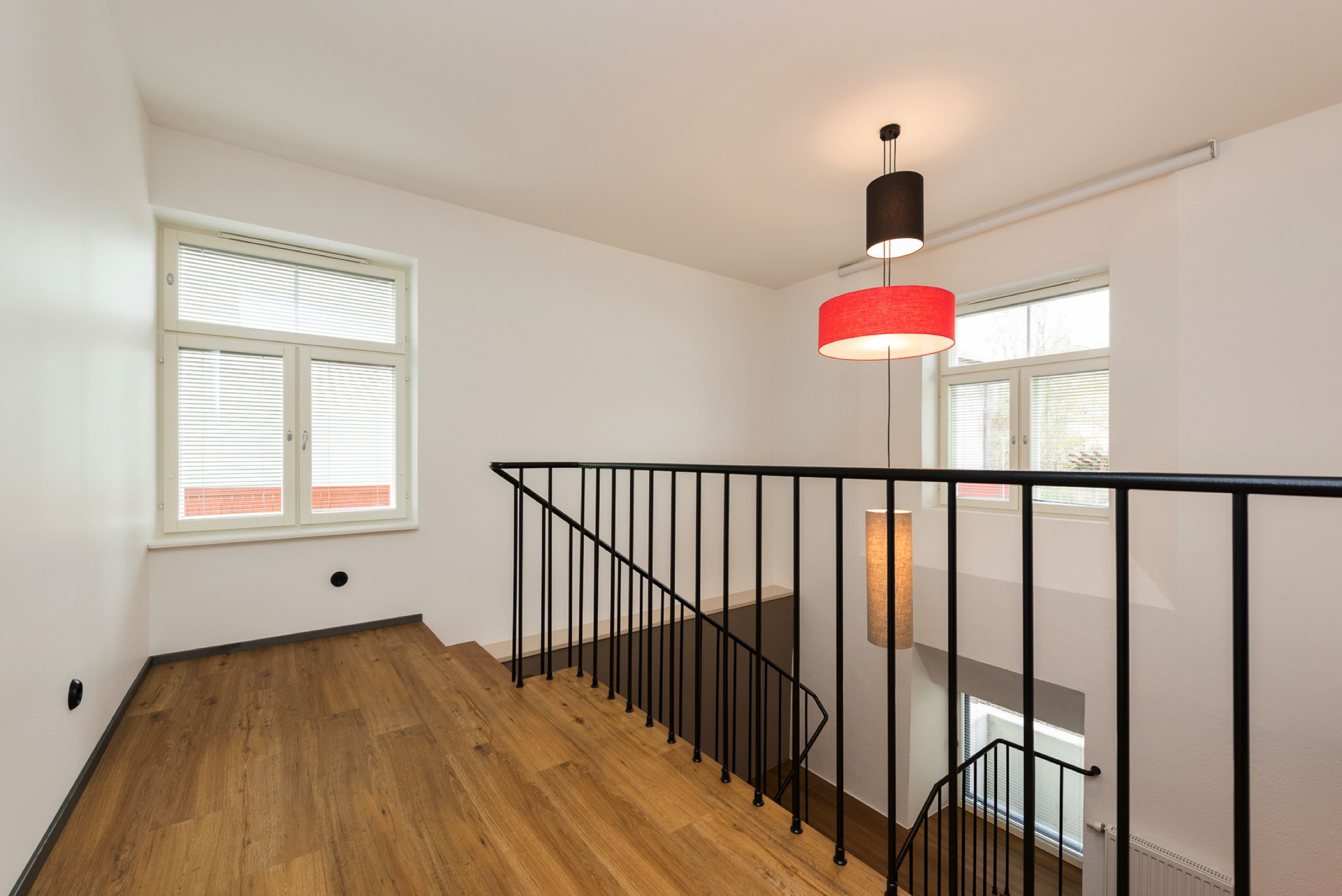 Una escalera con piso de madera y barandal negro, que lleva a una plataforma con una lámpara colgante roja.