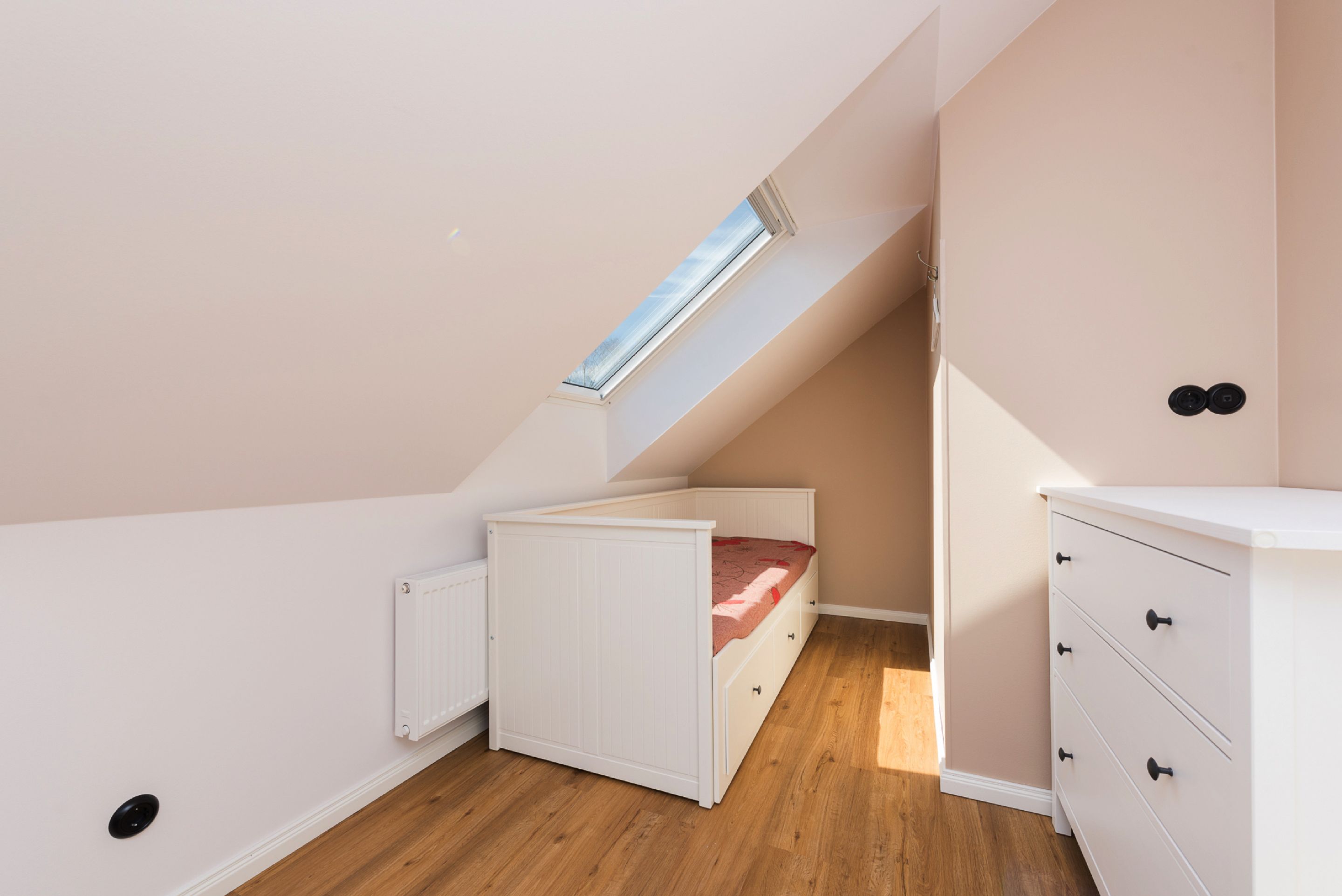 Una camera da letto con soffitto spiovente, una finestra a lucernario, un letto, una cassettiera e un termosifone.
